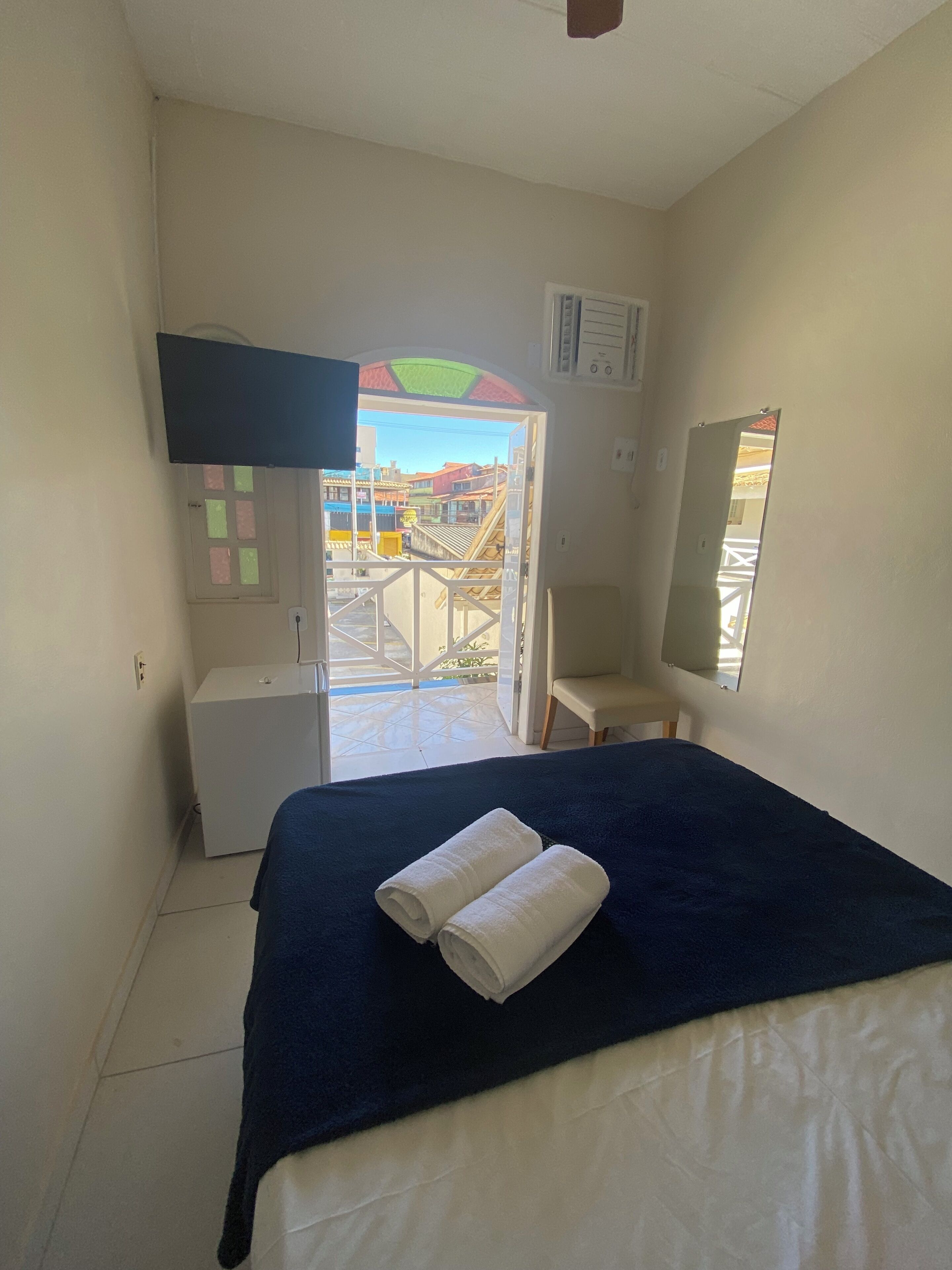 A hotel room with a bed, a desk, a chair, a TV, and a view of colorful buildings through an open door.
