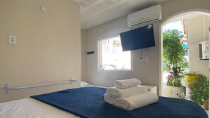 Una habitación de hotel con cama, ventilador de techo, aire acondicionado, televisor y ventana con vistas a edificios y plantas.