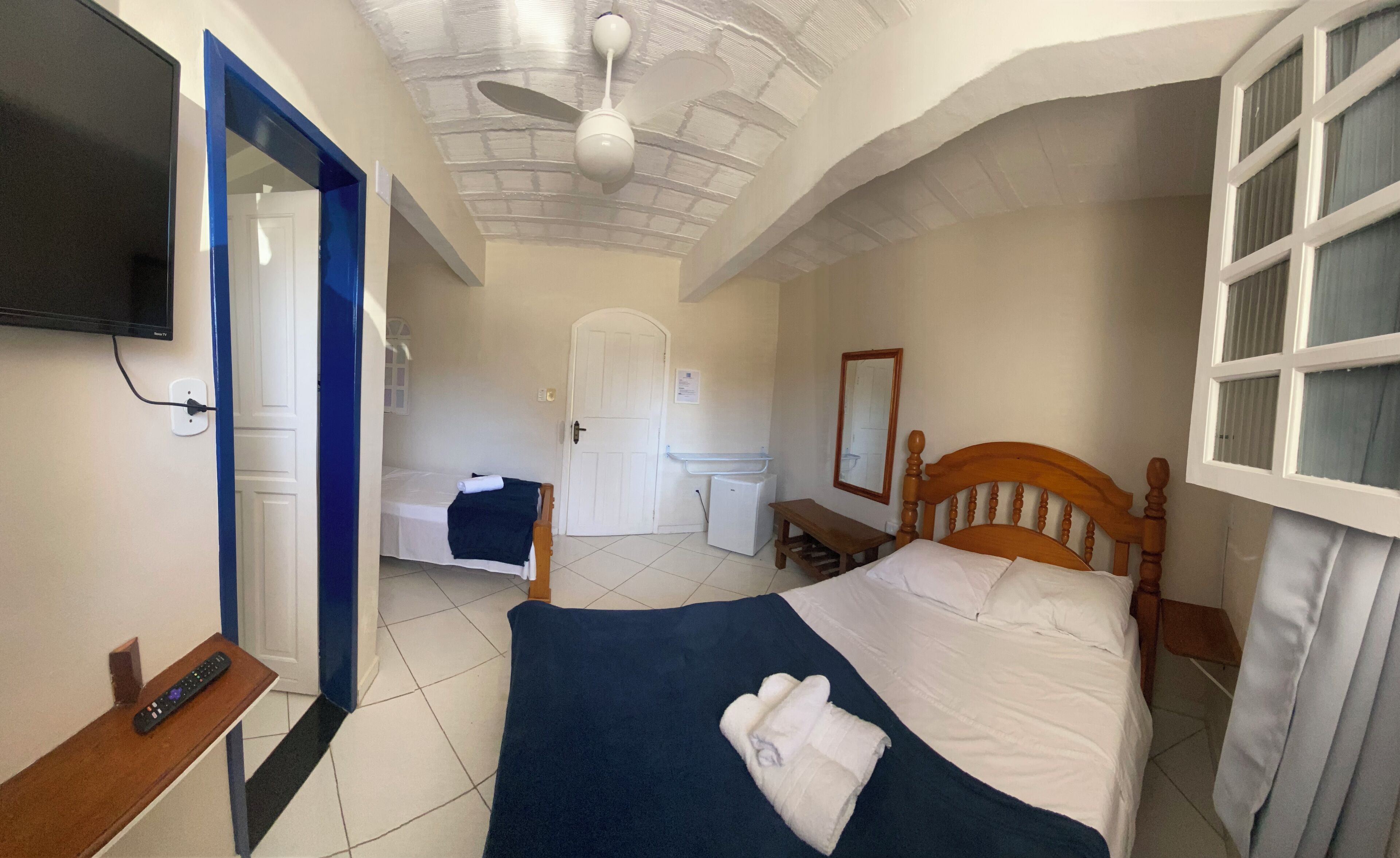 Una habitación de hotel con cama, escritorio, televisión, ventilador de techo y ventana.