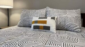 A neatly made bed with a geometric patterned bedspread and pillows, a bedside lamp, and a colorful abstract painting on the wall. - Divine Home Beach Retreat (Anavyssos)