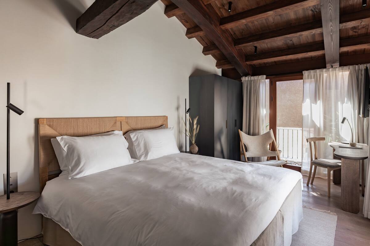 Un dormitorio con cabecera de madera, cama con ropa de cama blanca, mesita de noche, una silla, una pequeña mesa con lámpara y vistas al exterior a través de una ventana con cortinas.