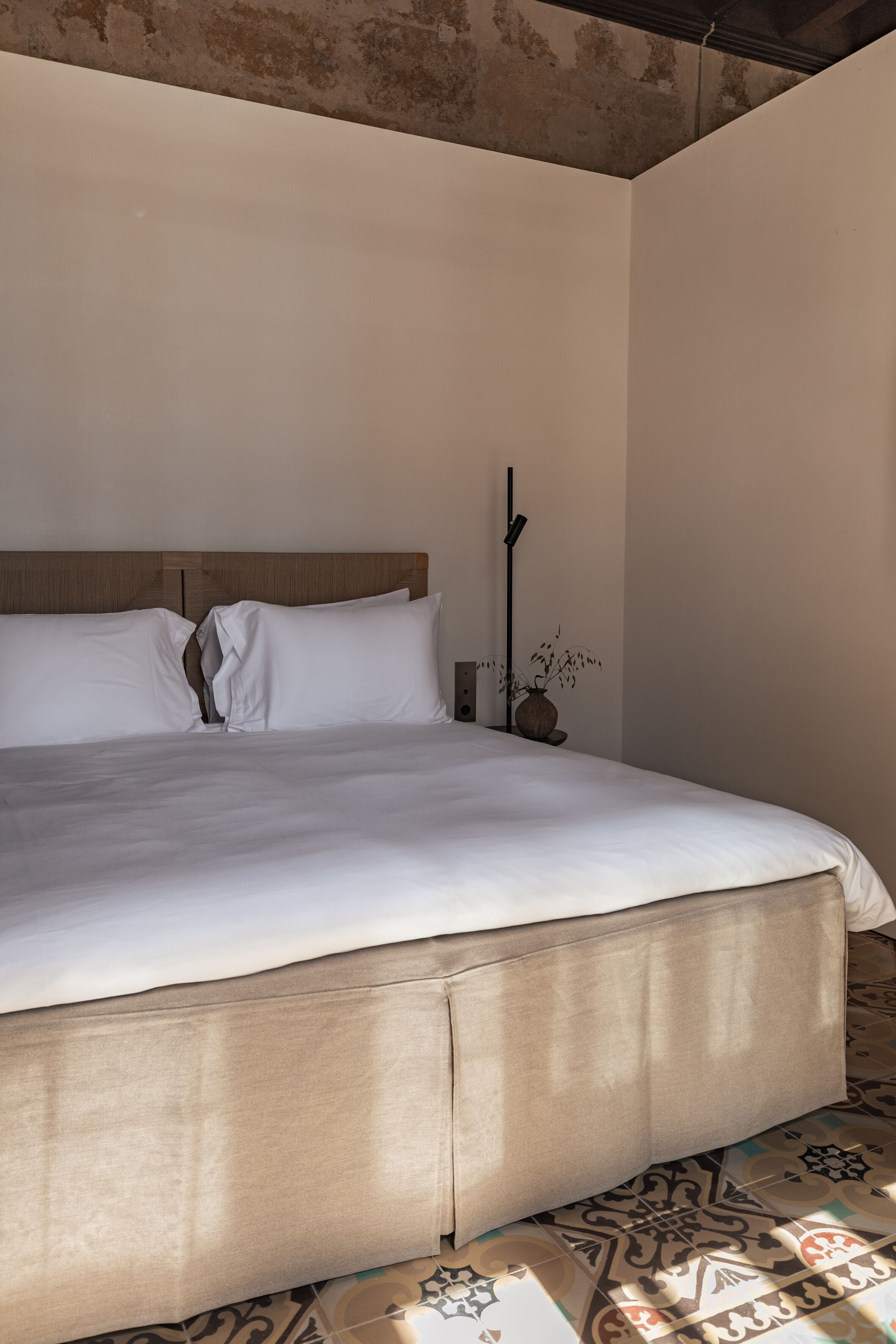 Una cama bien hecha con cabecera de madera, un jarrón con plantas secas y baldosas de suelo estampadas.