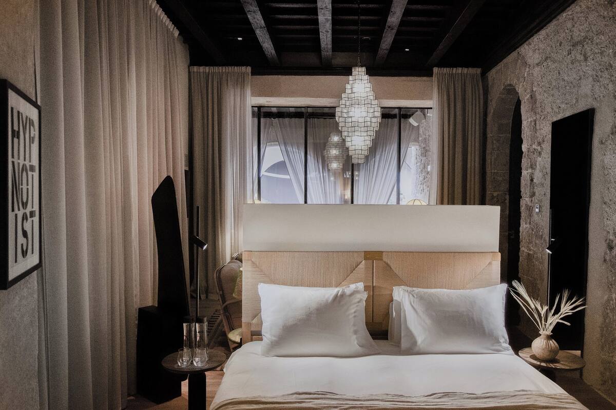 Habitación de hotel con una cama grande, un candelabro y vistas al exterior a través de una ventana.