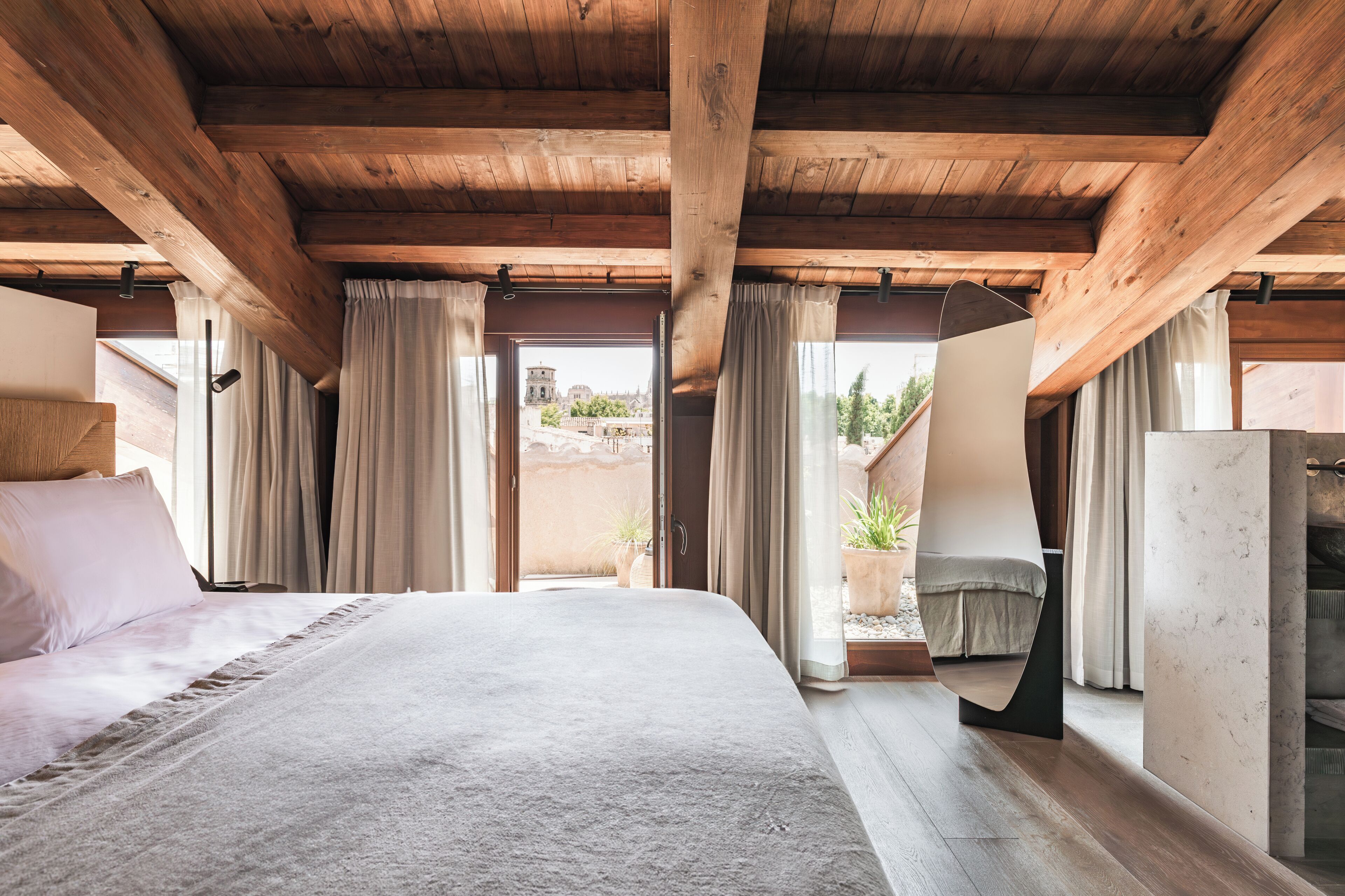 Ein Schlafzimmer mit einem großen Bett, Holzbalken an der Decke, Blick ins Freie und einem kleinen Balkon mit Pflanzen.