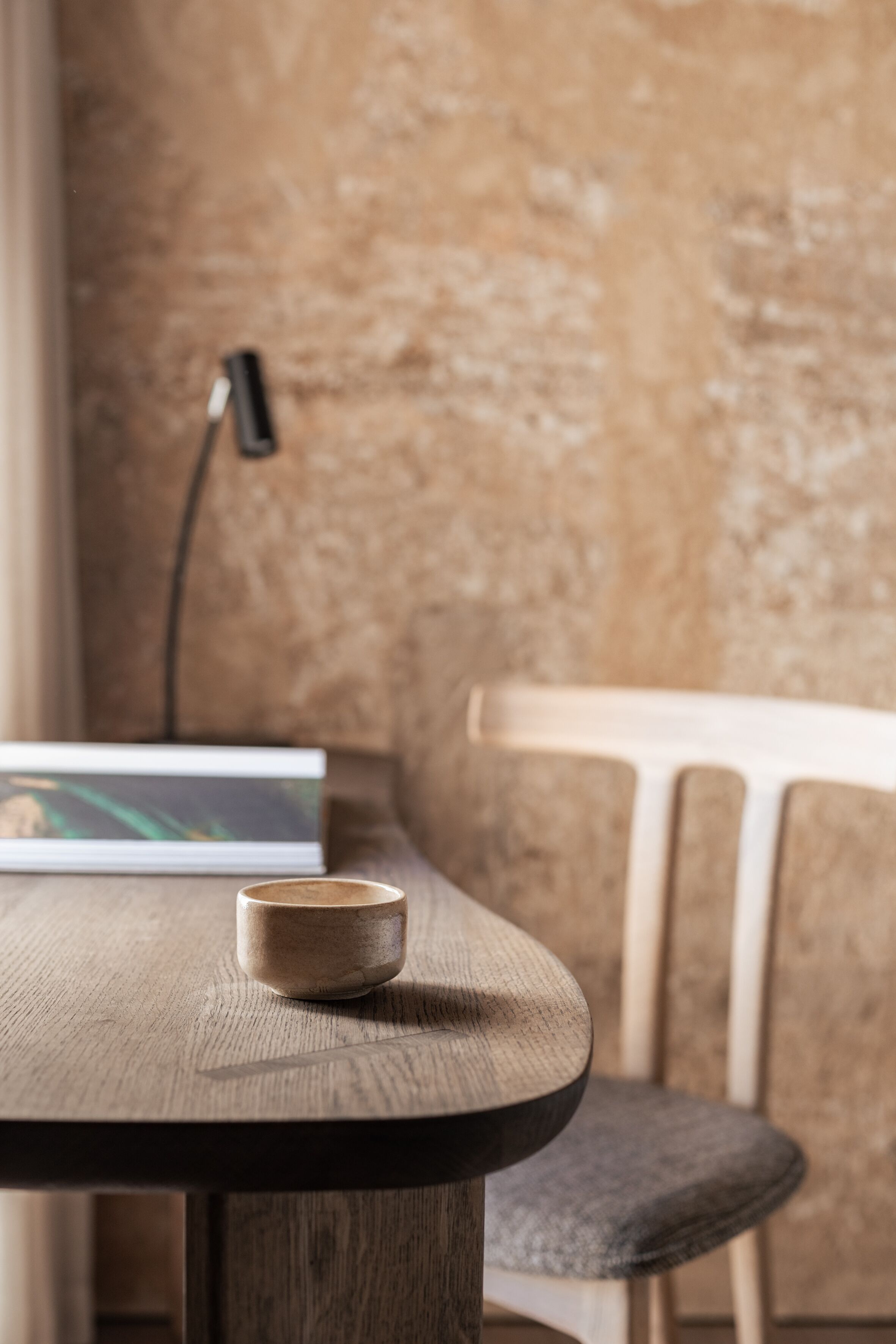 A wooden table with a cup, a laptop, and a chair in a room with a textured wall and a lamp.