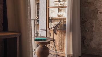 A room with a rustic wooden floor, a wicker chair with a book, and a view of a fountain through a window.