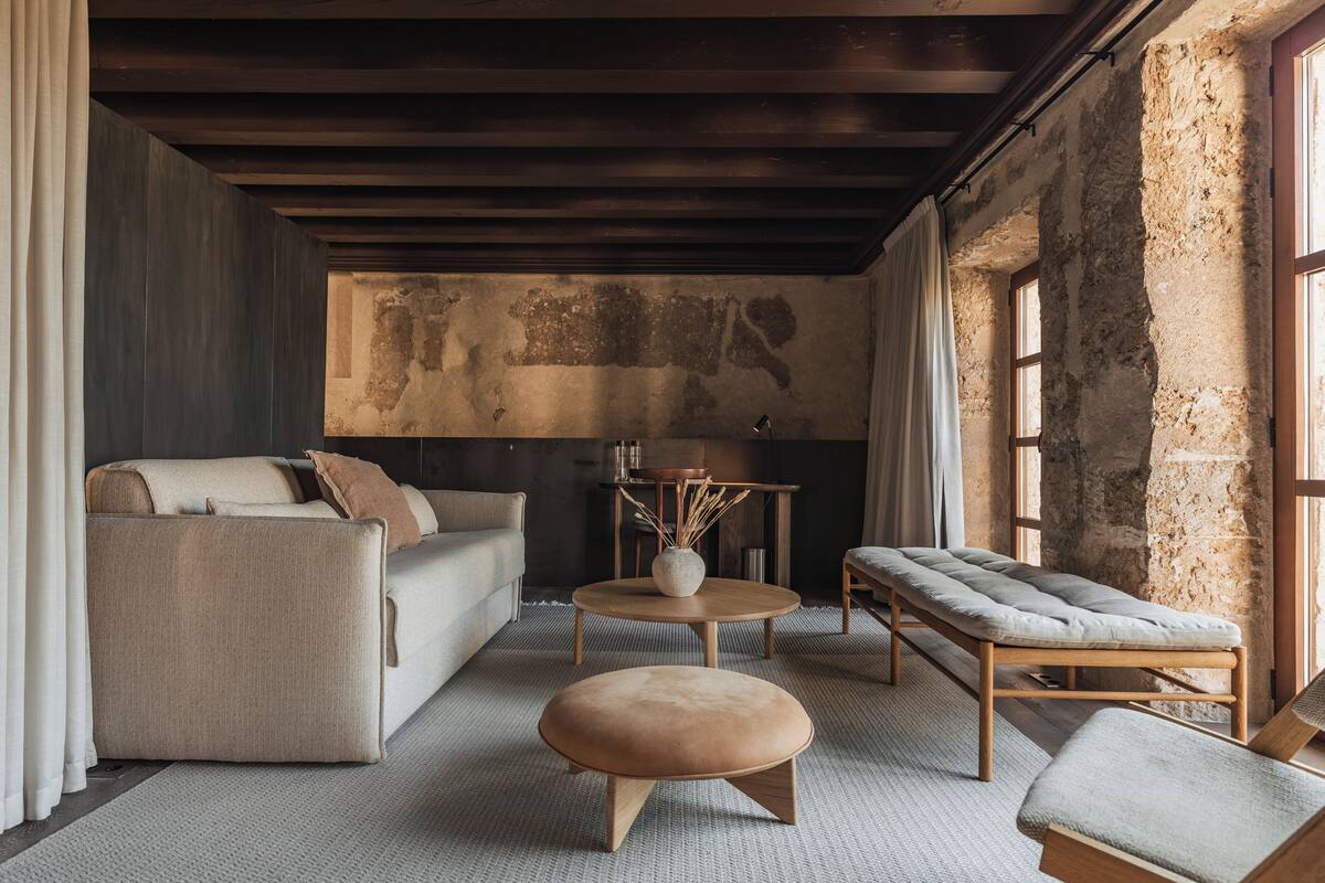 Una sala de estar acogedora con un sofá, reposapiés y un banco, una mesita con un jarrón, y un amplio ventanal con vistas al exterior.