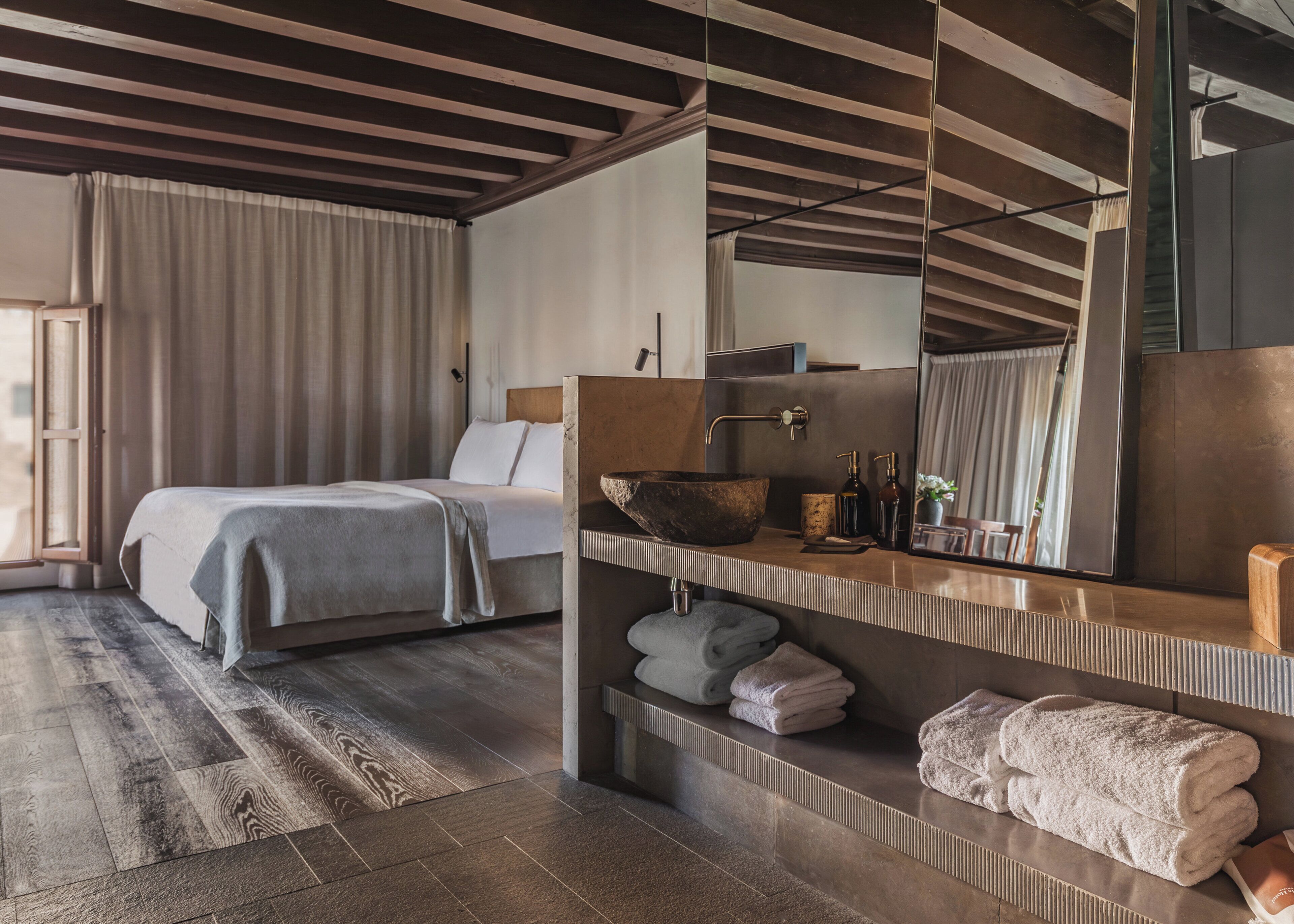 Ein modernes Hotelzimmer mit einem großen Bett, einer Holzbalkendecke, einem großen Spiegel und einem integrierten Waschbecken.