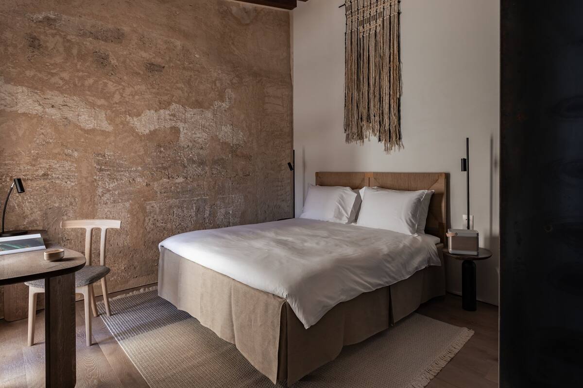Un dormitorio con una cama grande, un cabecero de madera, un macramé en la pared, una mesita de noche con una lámpara, y una pequeña mesa redonda con un libro y un cuenco.