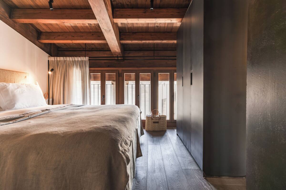 Un dormitorio con techo de madera, una cama con manta color beige, una mesita de noche y una cesta de mimbre.