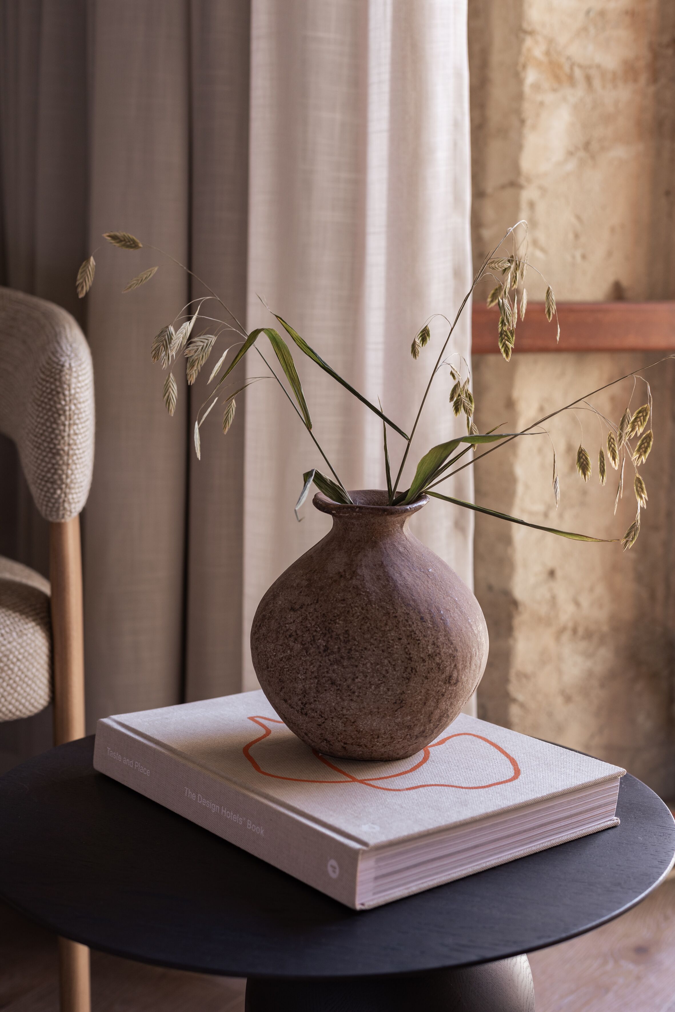 Un jarrón con plantas secas sobre una mesa, un libro titulado "The Design Hotels Book" y una silla con un cojín texturizado.