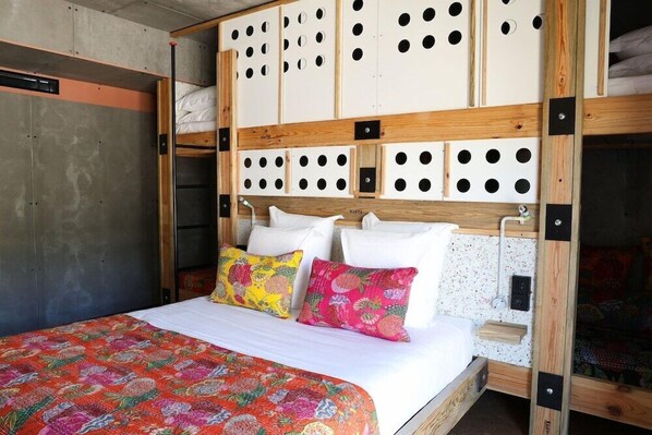Un lit recouvert d’une courtepointe aux couleurs vives, une tête de lit en bois ornée de motifs circulaires découpés, et un mur en béton.