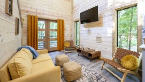 A wooden cabin interior with a sofa, ottomans, a wooden bench, a flat-screen TV, and large windows. - Pets Welcome! King BR+Loft w/Hot Tub/Lg Patio! (Hochatown)