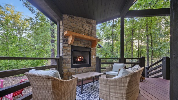 A covered outdoor seating area with wicker chairs, a stone fireplace, and a view of a forest. - Pets Welcome! King BR+Loft w/Hot Tub/Lg Patio! (Hochatown)
