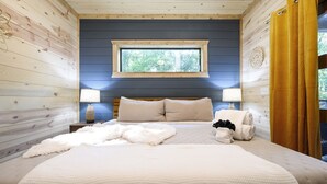 A bedroom with a wooden ceiling, a large bed with beige bedding, two bedside lamps, a window with a view of trees, and a yellow curtain. - Pets Welcome! King BR+Loft w/Hot Tub/Lg Patio! (Hochatown)