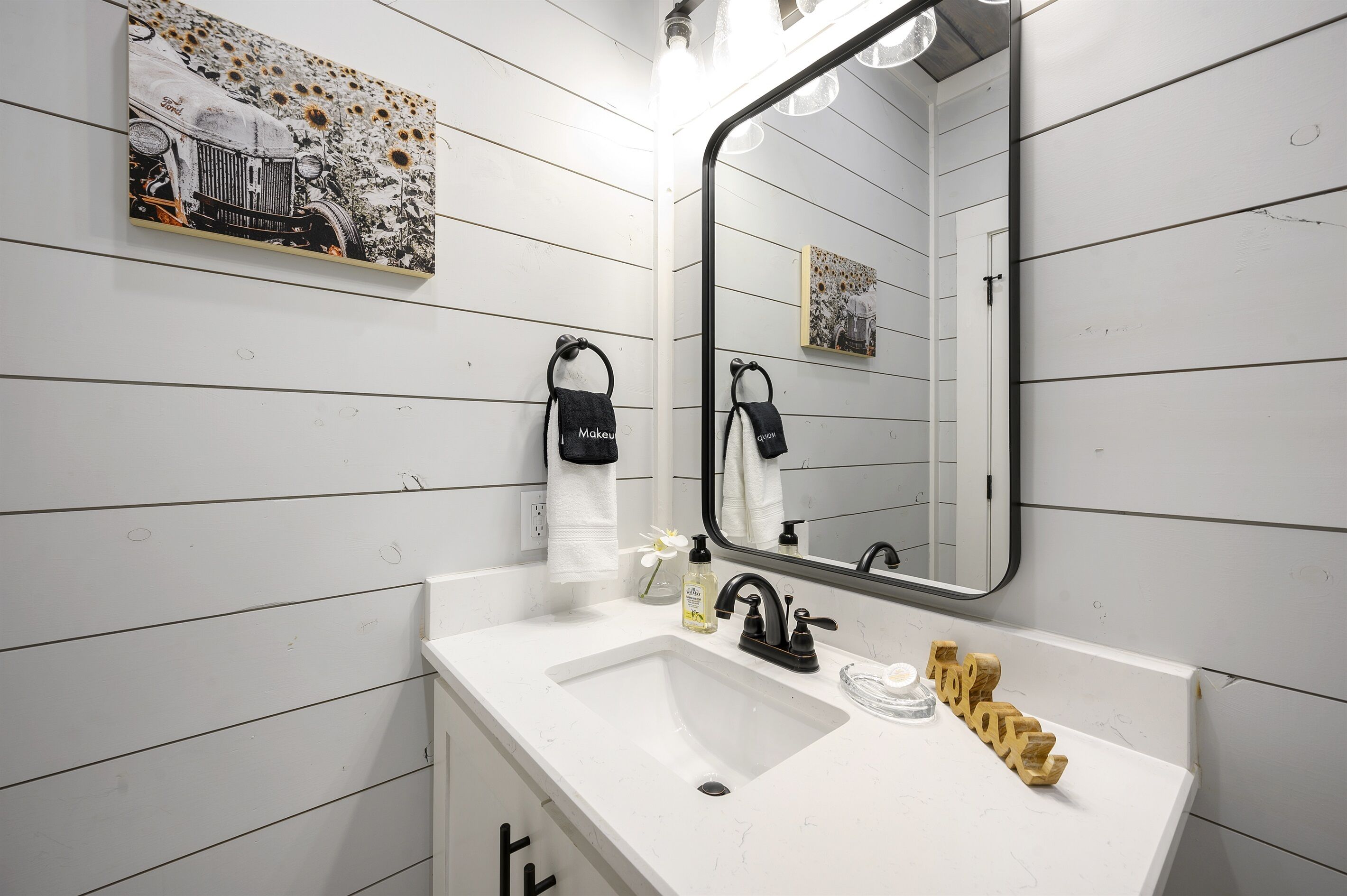 A bathroom with a white sink, black faucet, and a mirror above it. There are towels hanging on hooks and a small picture on the wall.