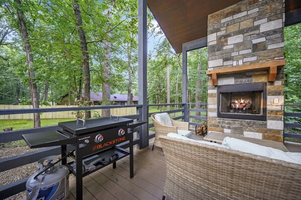 An outdoor deck with a stone fireplace, a grill, wicker furniture, and a view of a wooded area. - King BR+Gaming Loft, Hot Tub, Huge Patio! (Hochatown)