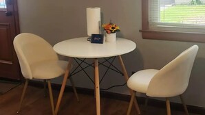Ein Zimmer mit einem runden Tisch, zwei Stühlen, einer kleinen Vase mit Blumen und einem Fenster mit Jalousien.