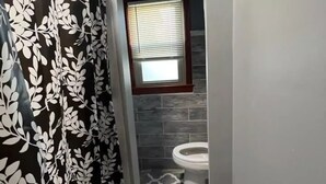 Ein schmales Badezimmer mit einer Duschvorhangs mit Blättermuster, einer Toilette und einem Fenster mit Jalousien.