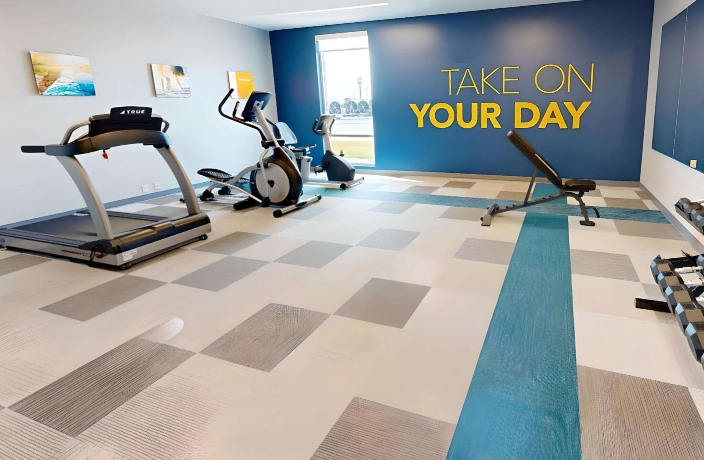 A gym with a treadmill, elliptical, and bench. The wall has a motivational quote.