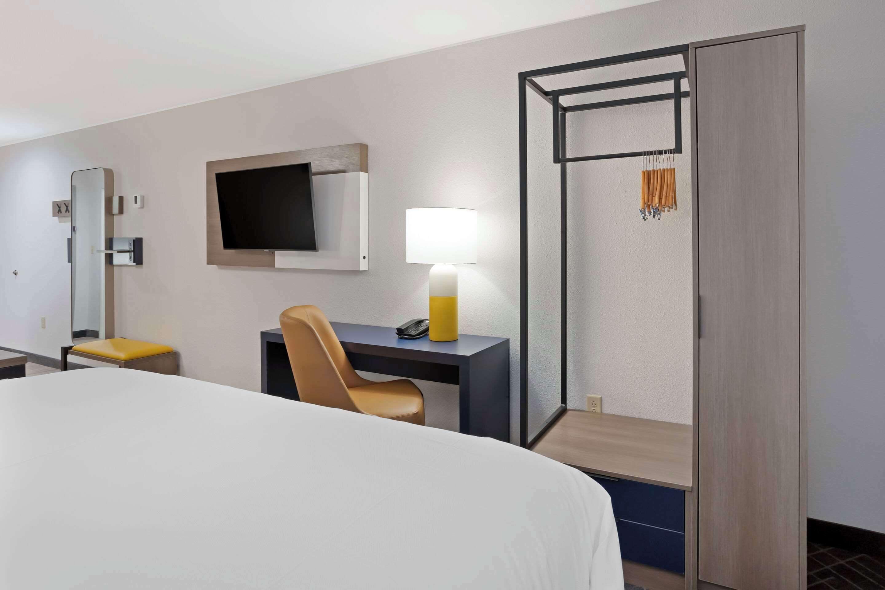 Una habitación de hotel con una cama, un escritorio con silla, un televisor montado en la pared, una lámpara y un armario.
