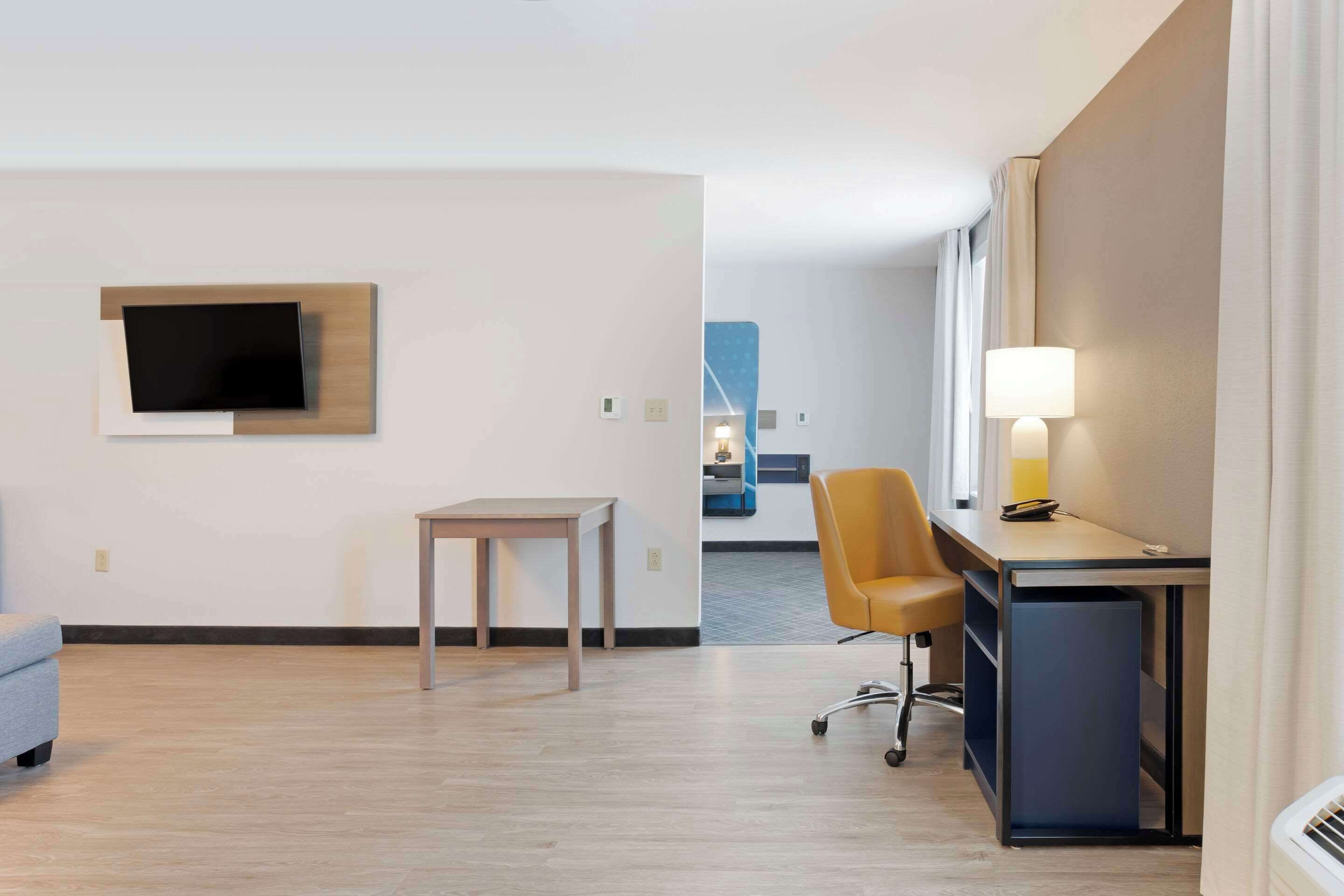 A hotel room with a desk, chair, TV, and a lamp.