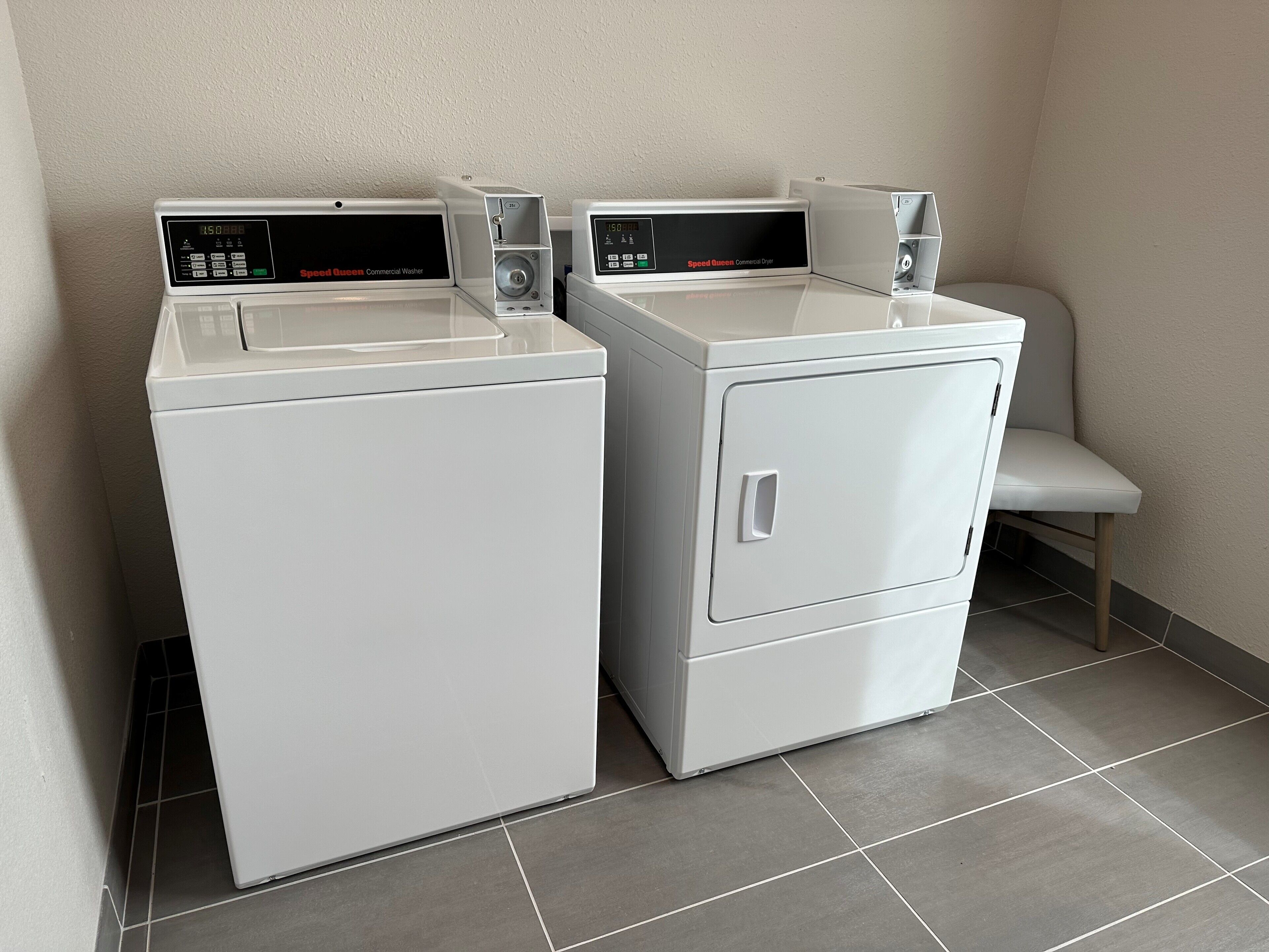 Twee witte wasmachines met digitale displays, een grijze betegelde vloer en een witte stoel in een hoek.