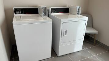 Two white front-loading washers with digital displays, a grey tiled floor, and a white chair in a corner.