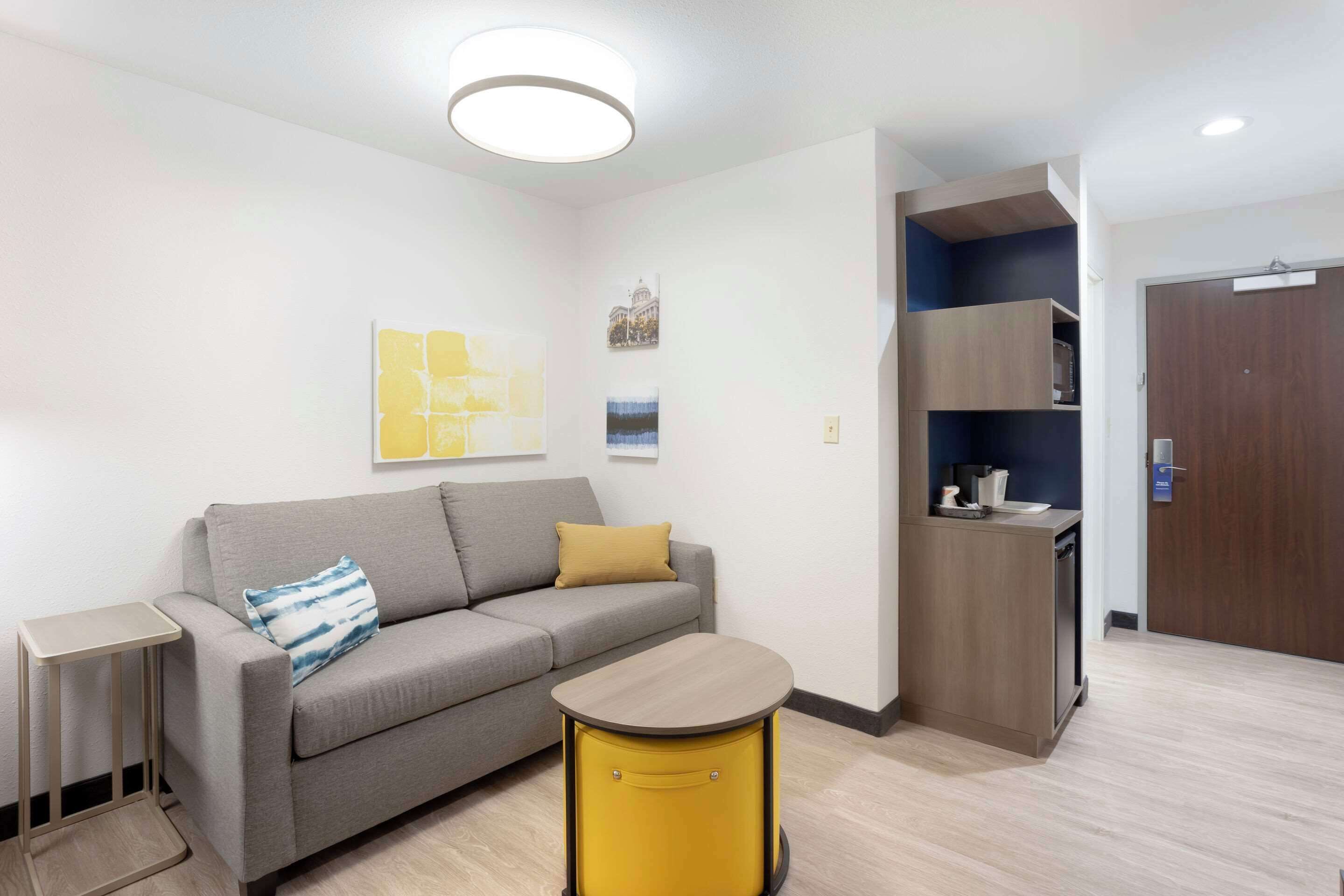 A modern living room with a grey sofa, a small round yellow table, and a kitchenette area with wooden cabinets and a microwave.