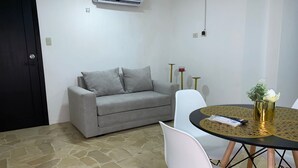 A room with a grey sofa, white chairs, a round table with a vase, and an air conditioning unit on the wall.