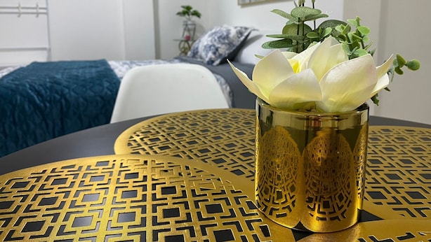 A room with a bed, a chair, a table with a decorative pattern, and a vase with a white flower.
