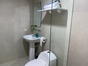 Une salle de bain avec un lavabo blanc, des toilettes et une étagère en verre au-dessus des toilettes, où sont rangées des serviettes.
