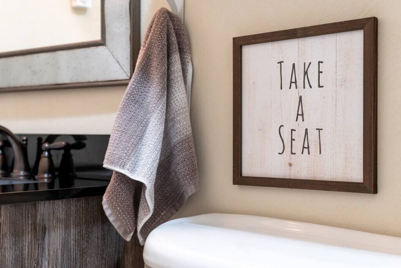 Une salle de bain avec un panneau encadré sur lequel on peut lire « ASSEOIEZ-VOUS » et une serviette accrochée au mur.