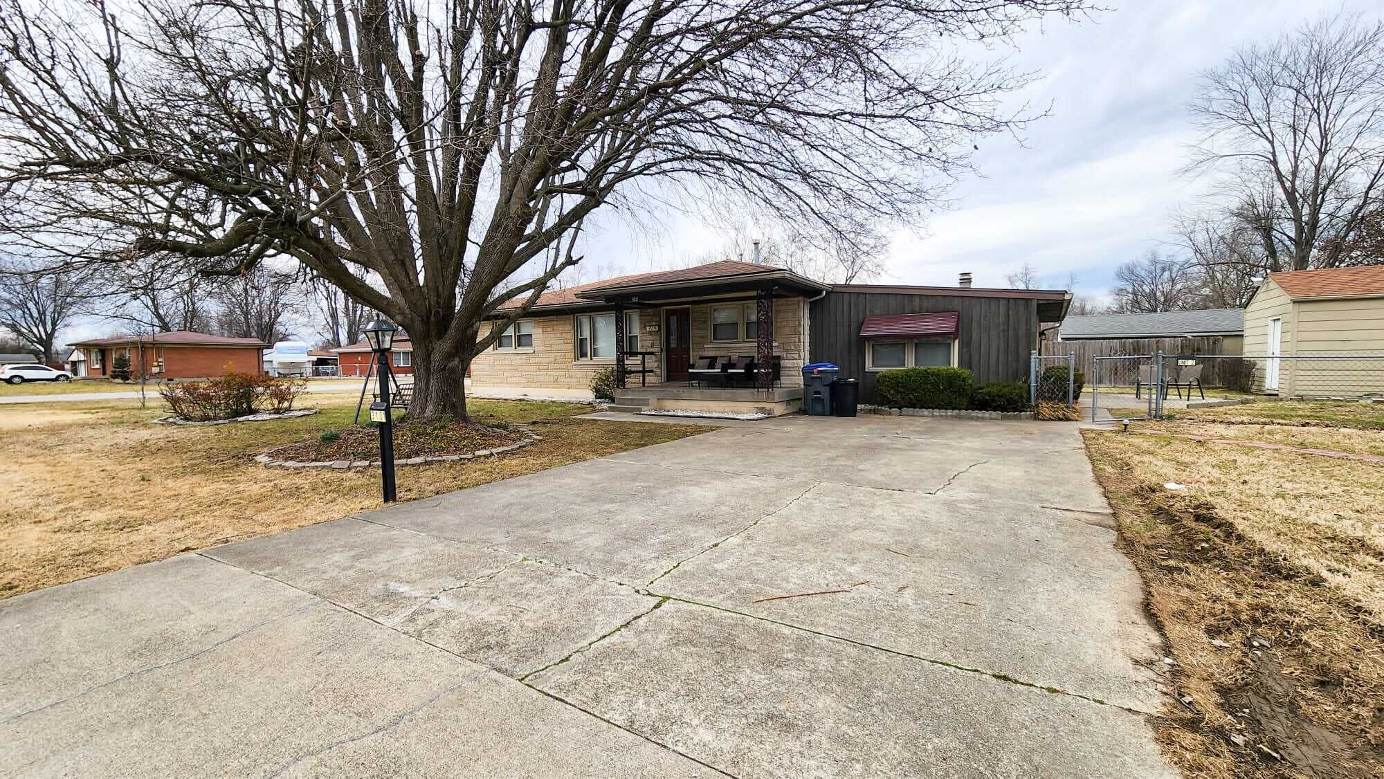 A residential house with a front porch, a driveway, and a large tree in the yard.