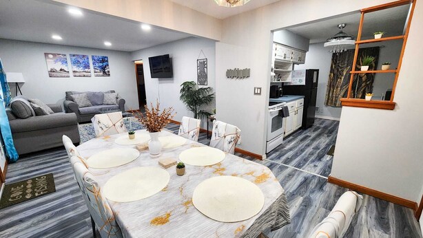 Ein modernes Wohnzimmer mit einem grauen Sofa, einem Esstisch mit Unterlagen und einer Küchenzeile inklusive Kühlschrank und Herd.