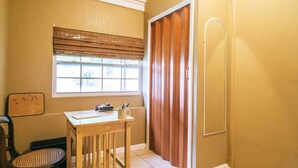 Een kleine kamer met een houten tafel, een stoel, een raam met zonwering en een deur met handdoekhouder.
