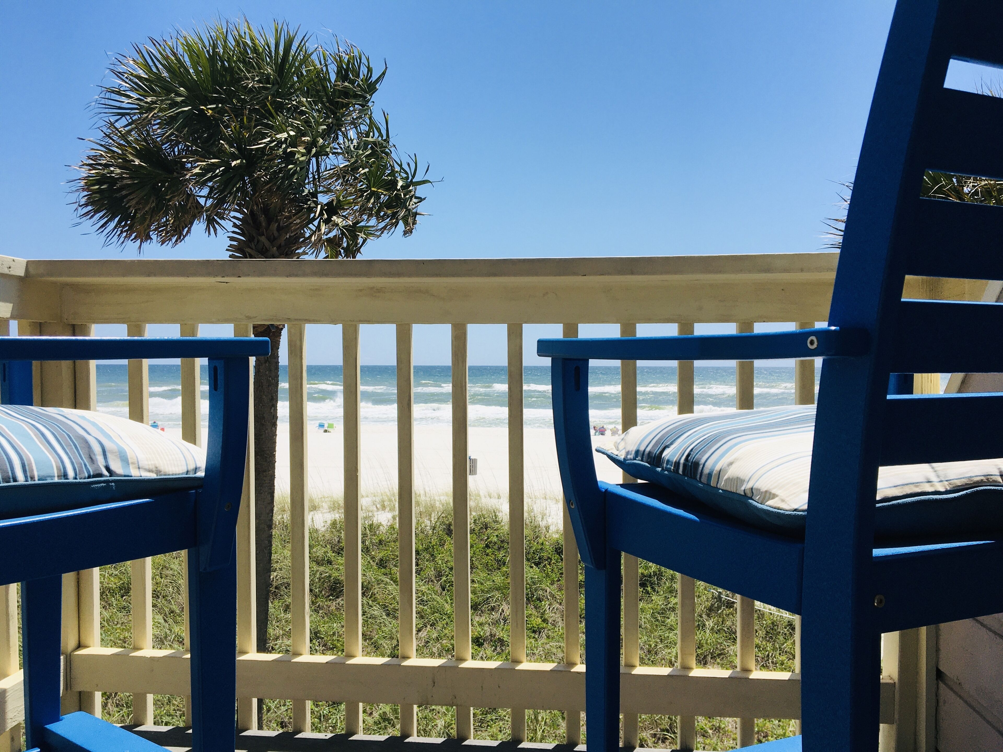 A balcony with two blue chairs and a palm tree, overlooking a beach with waves.