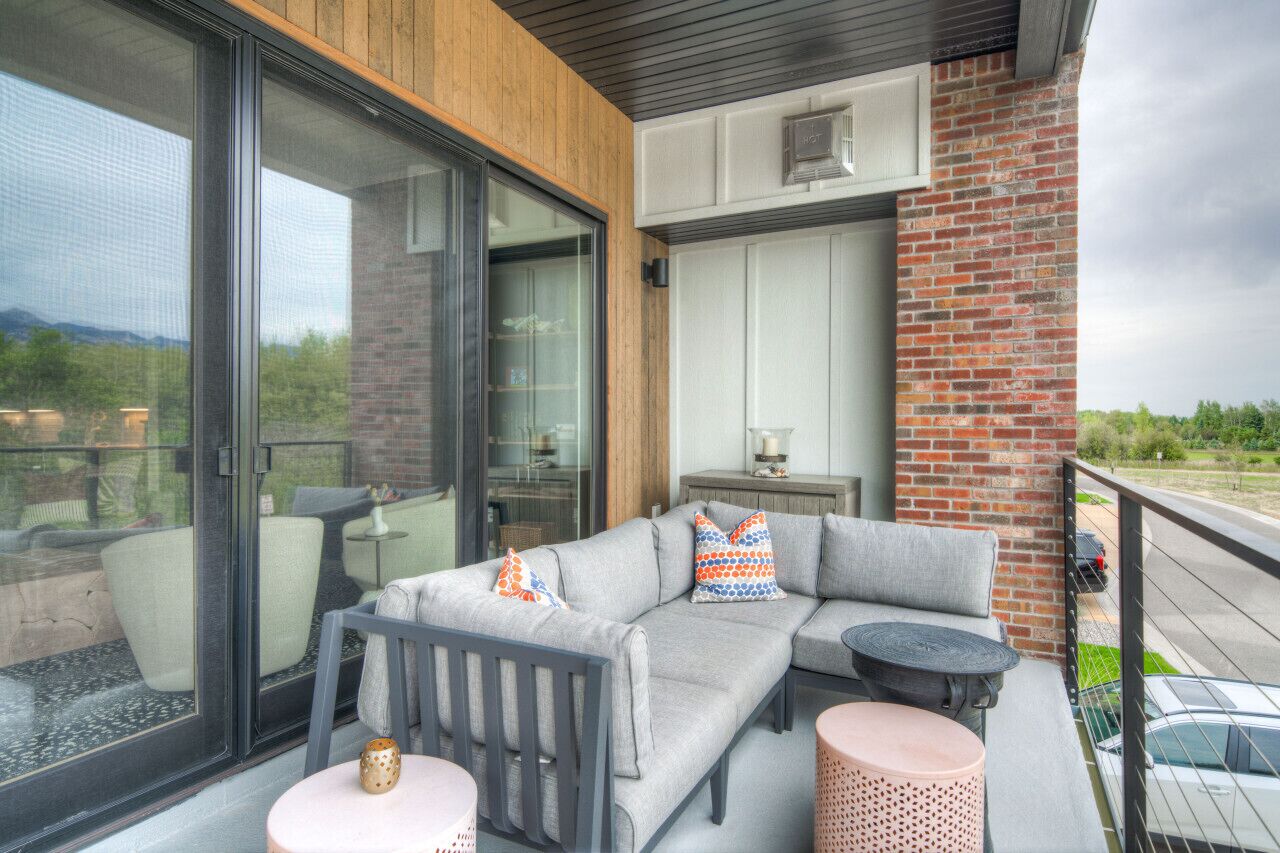 Ein Balkon mit einer grauen Eckcouch, einem kleinen runden Tisch und einem pinken Pouf.