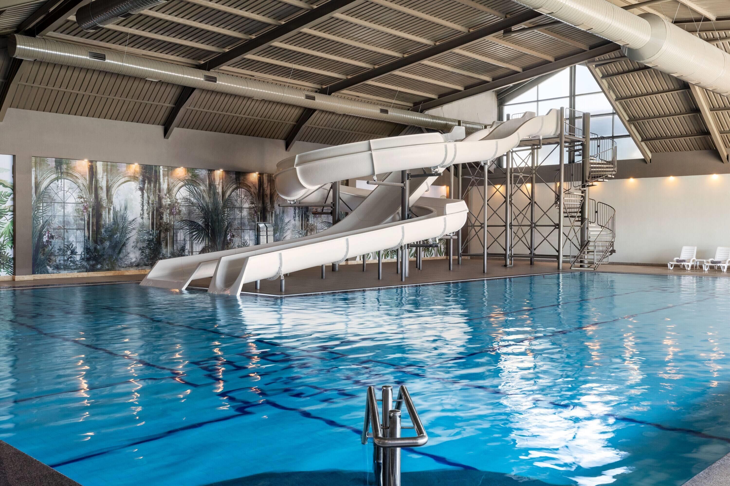 An indoor swimming pool with a water slide and a view of the sky through large windows.