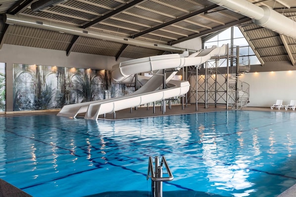 An indoor swimming pool with a water slide and a view of the sky through large windows.