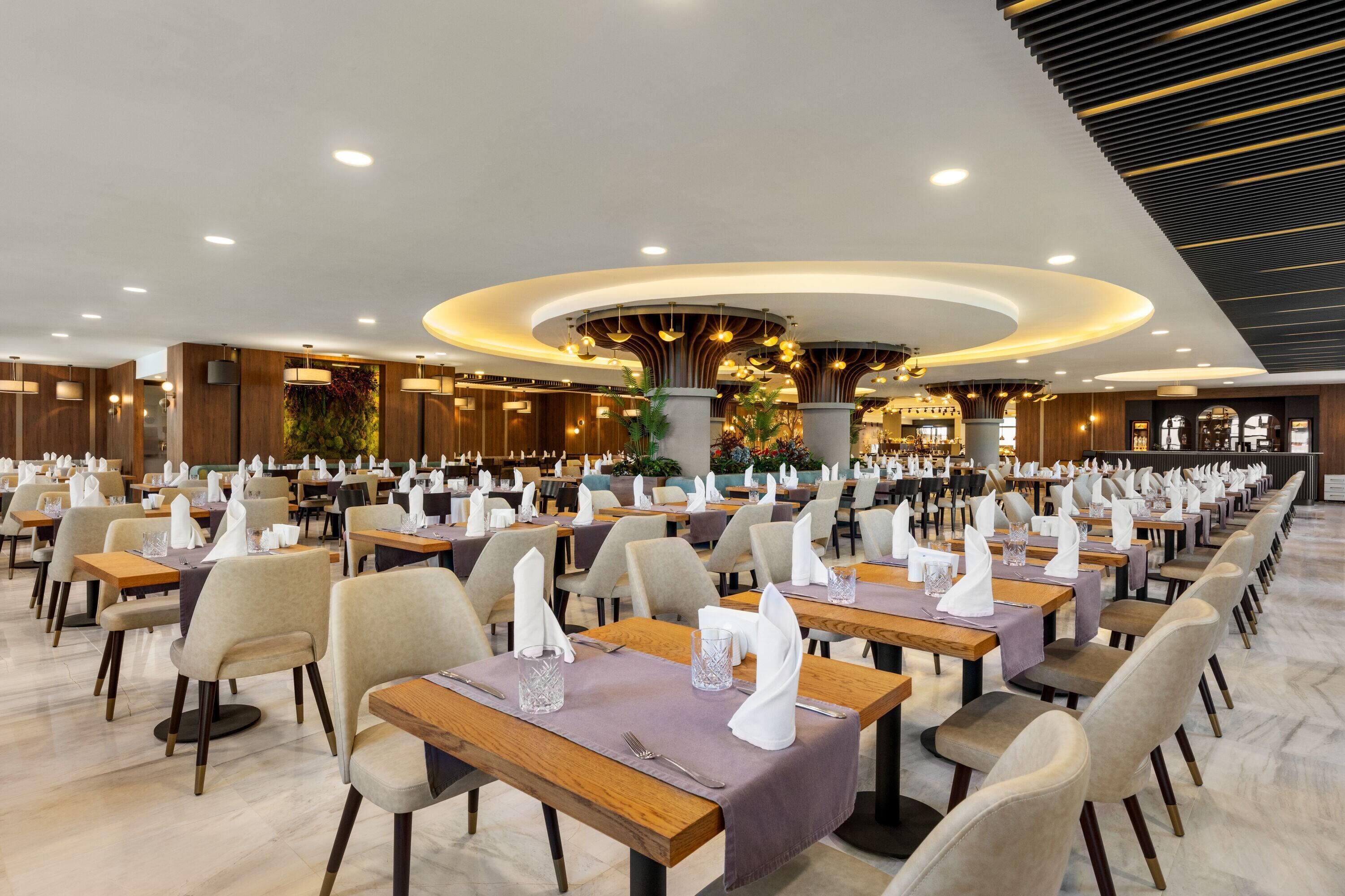 Um restaurante espaçoso com várias mesas e cadeiras, um detalhe decorativo central e um design moderno no teto.