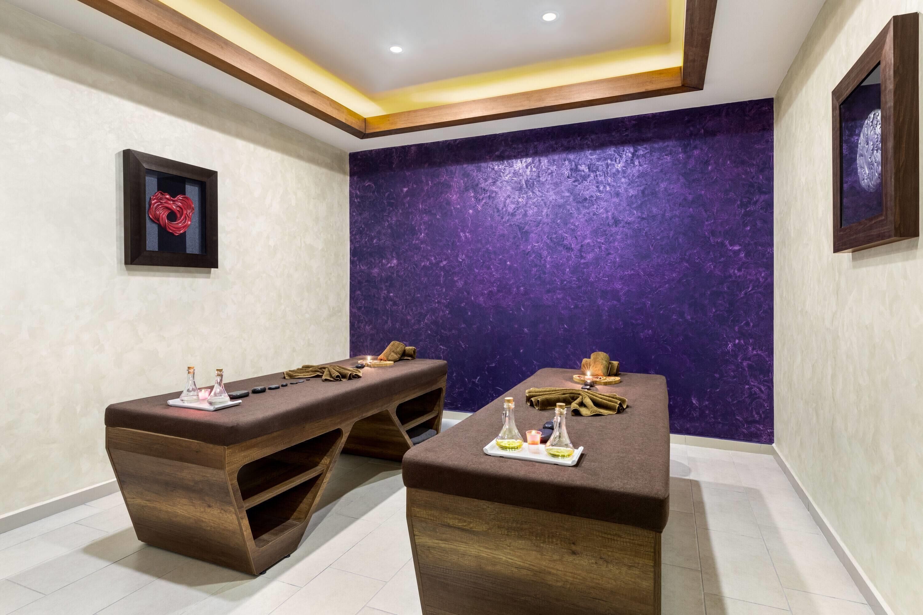 Una habitación con dos mesas de masaje de madera, una pared texturizada de color morado y una obra enmarcada a la izquierda.