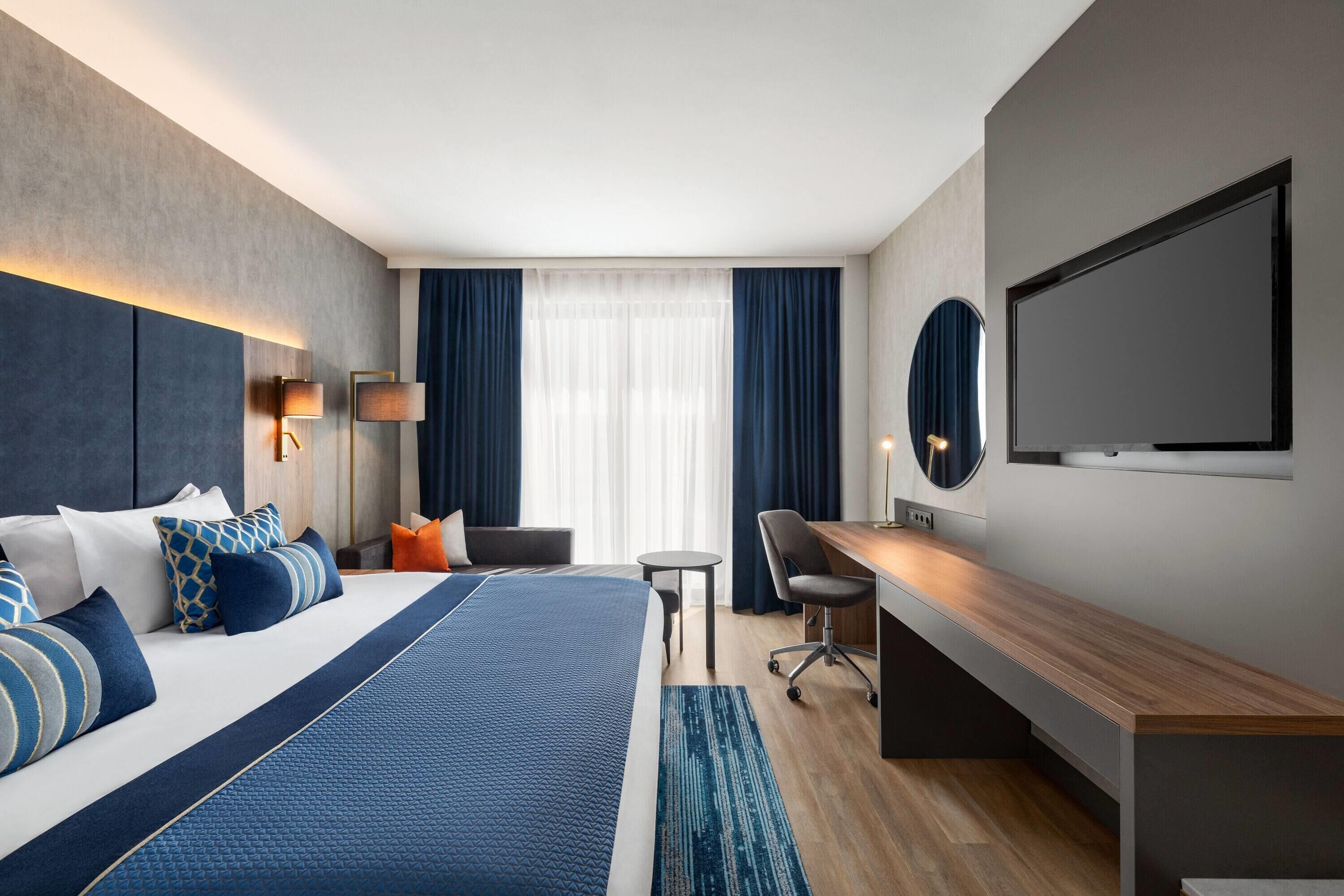 Habitació d'hotel moderna amb un llit gran, una taula de treball, una cadira, un televisor i una finestra amb cortines.