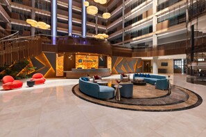 Un hall d’hôtel moderne doté d’un espace salon central, d’un comptoir d’accueil et d’un grand écran numérique.