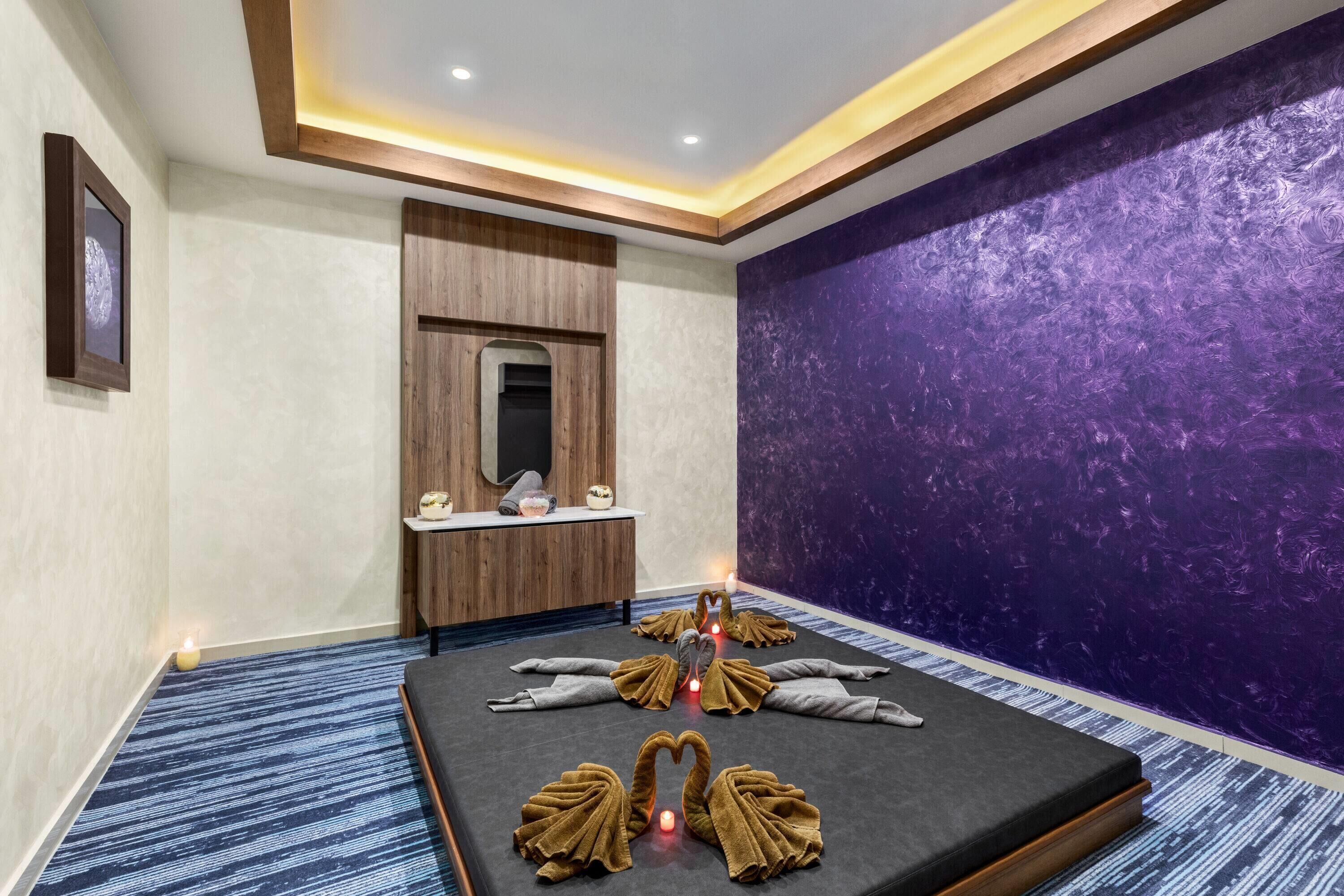 Et moderne spa-rum med et massagebord, håndklæder formet som svaner, et spejl og en væg med lilla struktur.