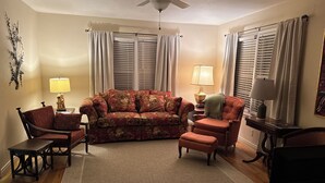A living room with a sofa, armchairs, a coffee table, and a rug. There are two lamps, a ceiling fan, and a wall-mounted decoration.