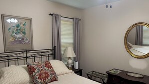 A neatly arranged bedroom with a bed, a bedside table with a lamp, a dresser, a mirror, and a framed artwork on the wall.