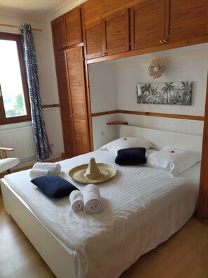 A neatly made bed with pillows, a hat, and towels, wooden cabinets, a window with curtains, and a wall art piece.