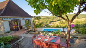A dining area with a table set for six, surrounded by red chairs, in a garden setting with a pool and a house in the background. - Dordogne gite, amazing views, private heated pool, large gardens, NO ADDED COSTS (Marnac)