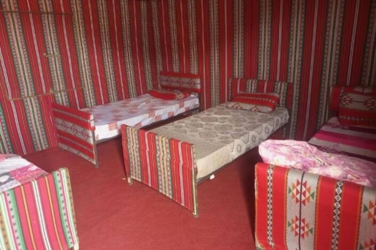 A room with two single beds, each with a patterned bedspread and pillows, and a small table with a tablecloth. The room has striped wallpaper and a red carpet.